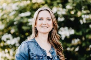 Kat Foxx Photo Kathleen Foxx, a white woman with blue eyes and long, dark blonde, wavy hair. She's smiling with teeth and wearing a denim jacket, standing in front of a green bush with white flowers, which is blurred in the background.