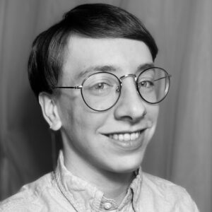 Dougherty_Editorial Brochure Photo A photo of Oliver Dougherty, a white androgynous person wearing round glasses, a button-down, and a single dangling earring