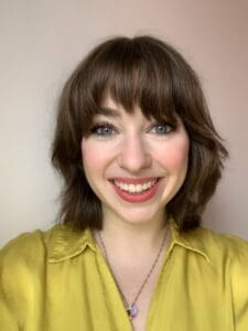 Clara Foster Agent Headshot of a white woman with shoulder-length brown hair and a fringe smiling directly into the camera. She's wearing a light green silky shirt and has pink lipstick and rosy cheeks.