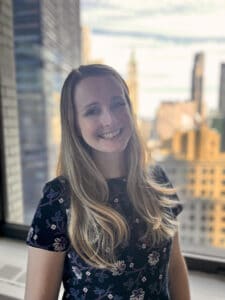 website photo Smiling white woman in an office. Downtown New York is visible through the window behind her.